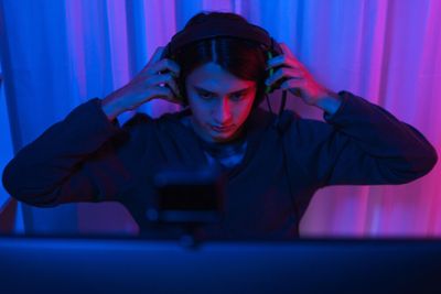 A young man places his headset over his head, while seated in front of a computer screen, in a room lit with LEDs.