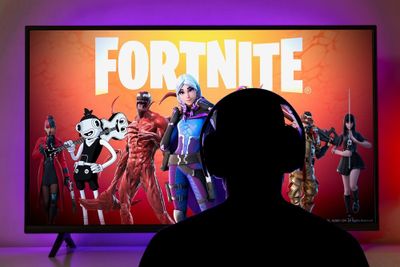 The silhouette of person in front of a large LCD screen, displaying the Fortnite logo along with various in-game characters, against a red background.