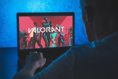 A PC gamer seated with his laptop open, with the screen displaying Valorant characters against a red background, with the game's name displayed in the forefront.