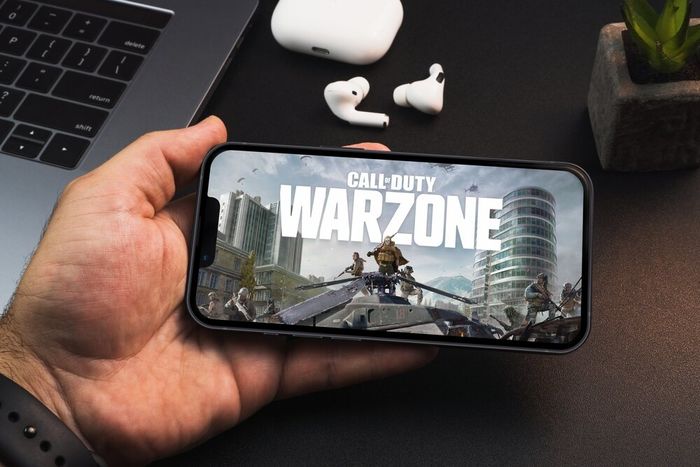 A person holding their cellphone, displaying the Call of Duty: Warzone opening page, with a laptop, wireless earphones, and small plant visible on the desk.