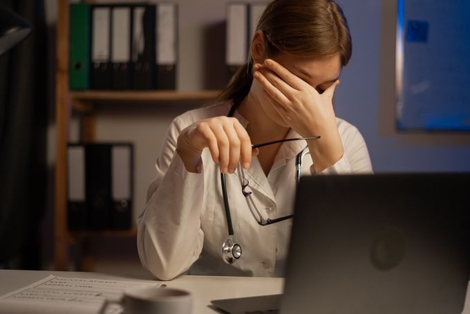 A woman with a stethoscope sitting at a desk in front of a laptop holding her head in her hand.