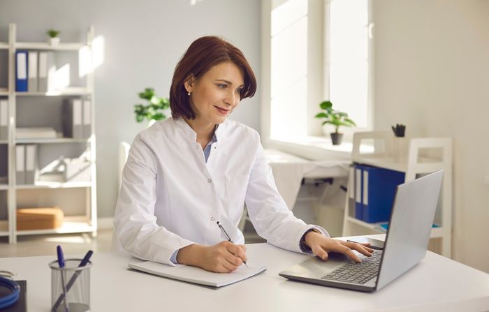 A female doctor sitting at a desk working on a laptop whle tang notes.