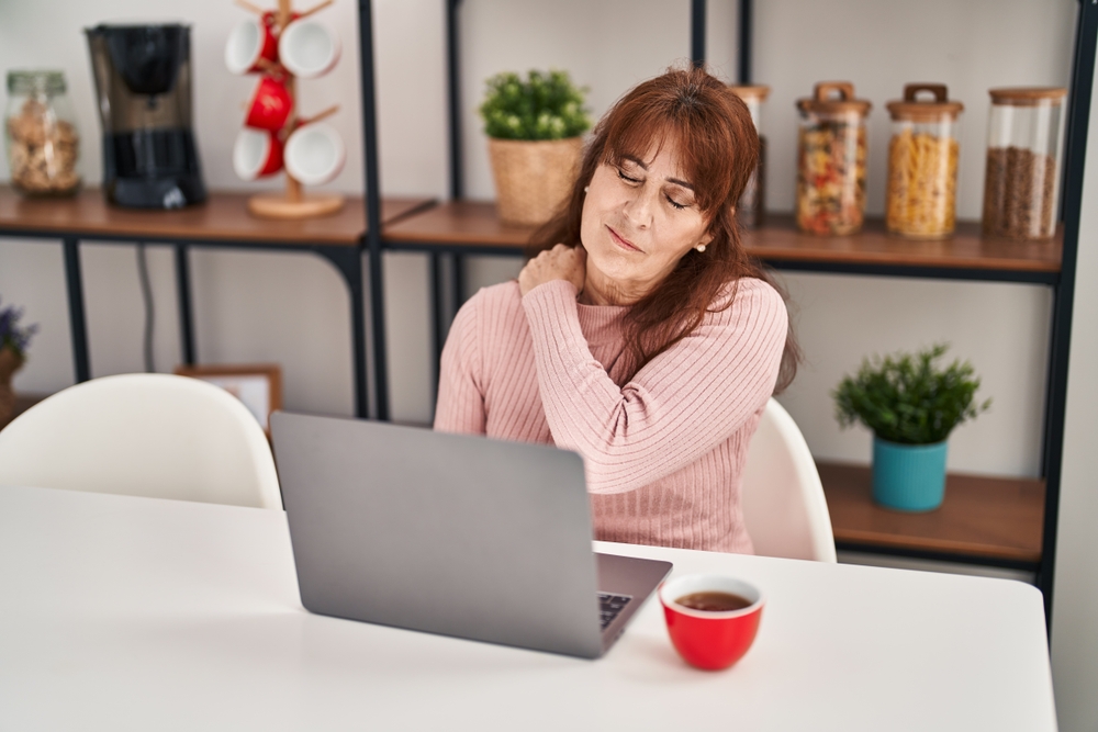 Elderly woman sitting at table and rubbing her inflamed shoulder due to