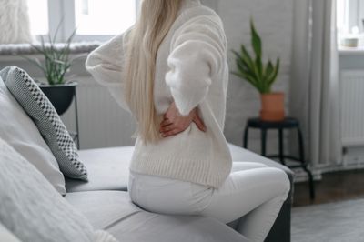 Woman in white clothing sitting on her couch rubbing her lower back due to pain