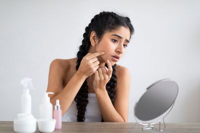 Woman closely examining skin blemishes in a small mirror next to various anti-ageing skincare products
