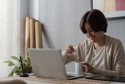 Smiling woman sitting in front of laptop while pouring CBD oil into cup