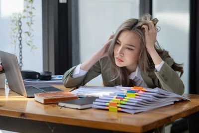 Physically and emotionally stressed businesswoman sitting at work desk in front of piles of papers