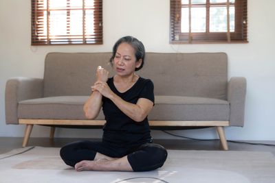 Elderly woman sitting cross-legged on the floor massaging wrists while dealing with menopause joint pain