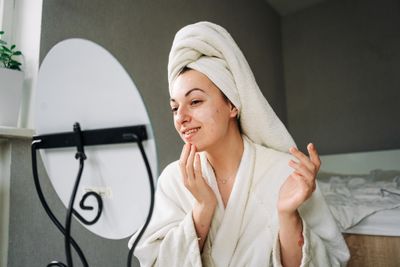 Woman in robe with towel on her head examining her face skin in mirror