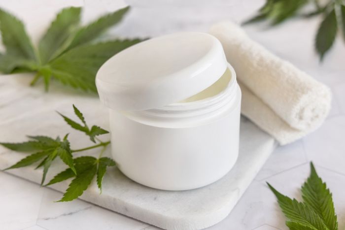 A jar of CBD moisturising cream sitting on top of a white counter surrounded by cannabis leaves