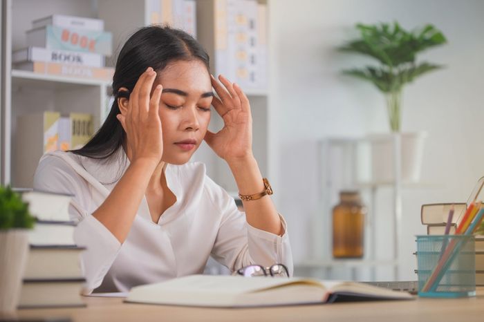Stressed woman sitting at her work desk surrounded by books with her eyes closed and rubbing her temples
