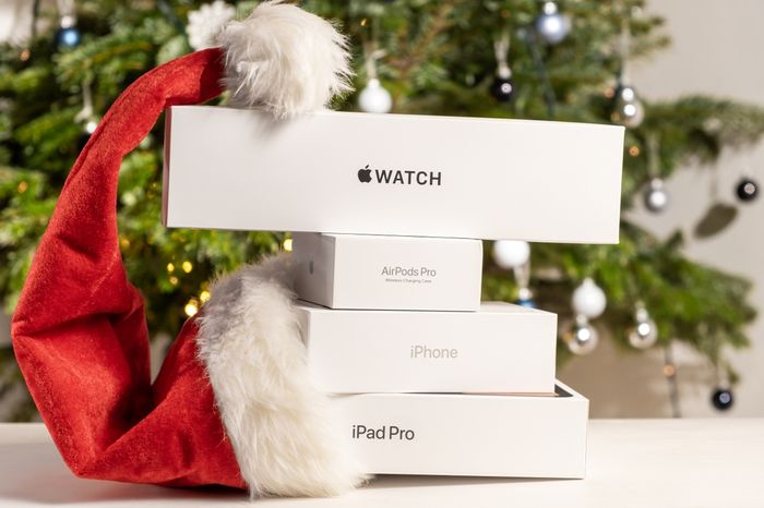 Various unopened Apple products stacked in front of a Christmas tree next to a Santa hat
