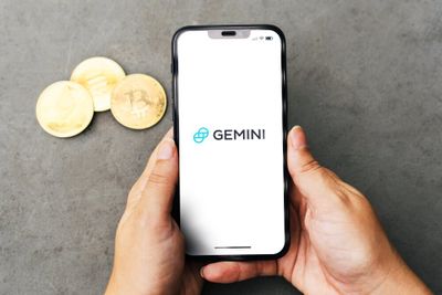 A woman's hands holding a smart phone, with the Gemini logo displayed against a white background, with cryptocurrency coins visible on the table top.