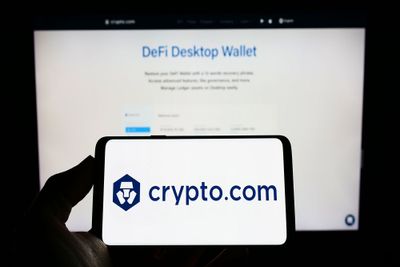 A person holding a smart phone, displaying the Crypto.com logo against a white background, with  a laptop screen displaying their DeFi Desktop Wallet landing page visible in the background.