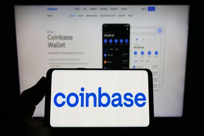 A person holding their smartphone, displaying the Coinbase logo in blue against a white background, with the Coinbase Wallet web landing page visible on a computer screen in the background.
