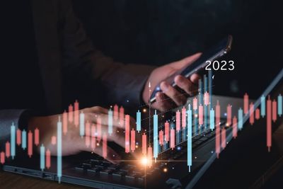 A man holding a phone in one hand while typing on a laptop keyboard, featuring stock market graph overlay  for 2023.