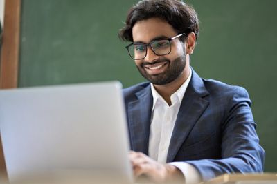 A young man with professional attire, spectacles, and a pleasant facial expression, busy working on his laptop.