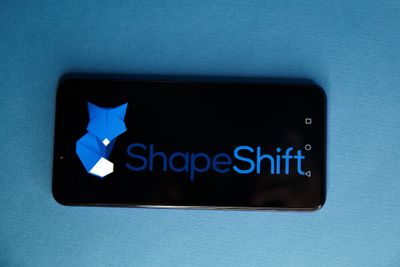 ShapeShift's name and fox-logo in blue displayed on a black phone screen, resting against a blue background.