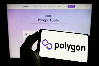 A smartphone displaying the Polygon logo against a white screen, with the Polygon website open on a computer screen visible in the back.