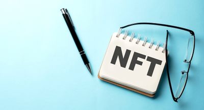 NFT written on a notepad with a pen and glasses alongside on a light blue background