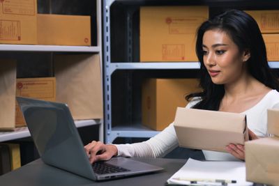 A woman seated at a desk in a warehouse office, inputting values into her laptop, while holding a box in her left hand.