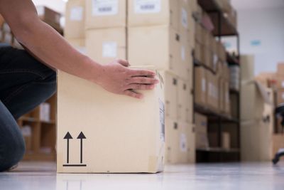 A person about to pick a box, marked as fragile, up from the floor of their warehouse, with various shelves containing other boxes visible in the background.