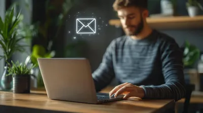 a man sitting at a table using a laptop, there's an email icon floating above the laptop