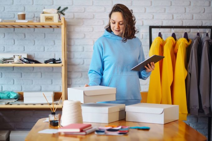 A woman in a blue sweater holding a tablet while looking through packages stacked on a desk