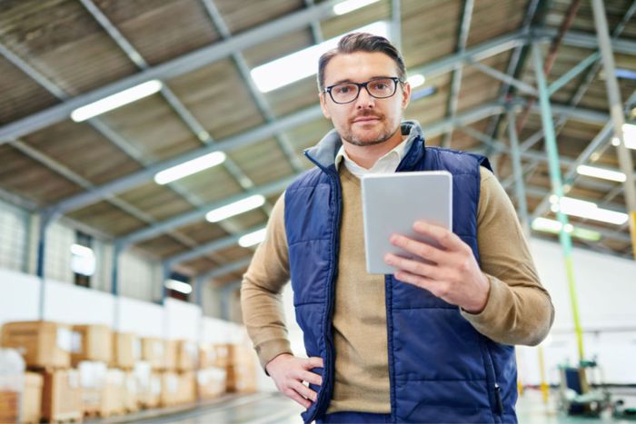 A professional standing in a warehouse, holding a tablet, with his other hand placed on his hip.