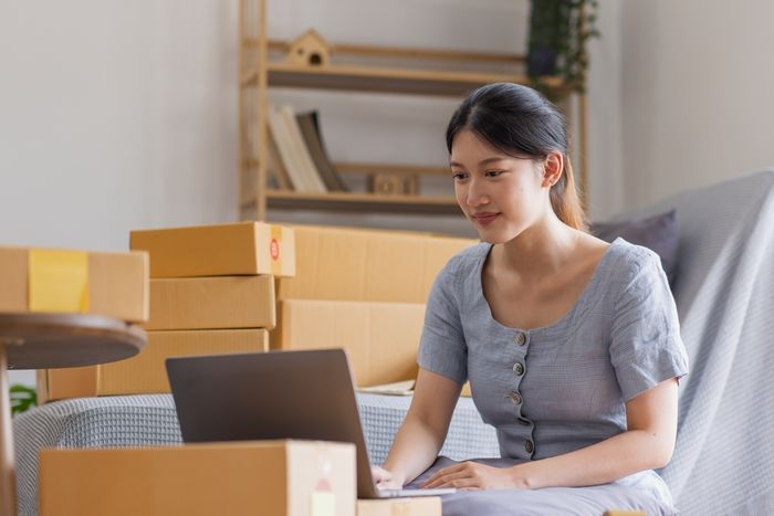 A woman seated in her living room, working on her laptop, with various boxes visible in the background.