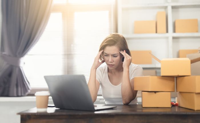 Shopify seller sitting in front of a laptop computer stressed and holding her head surrounded by deadstock boxes