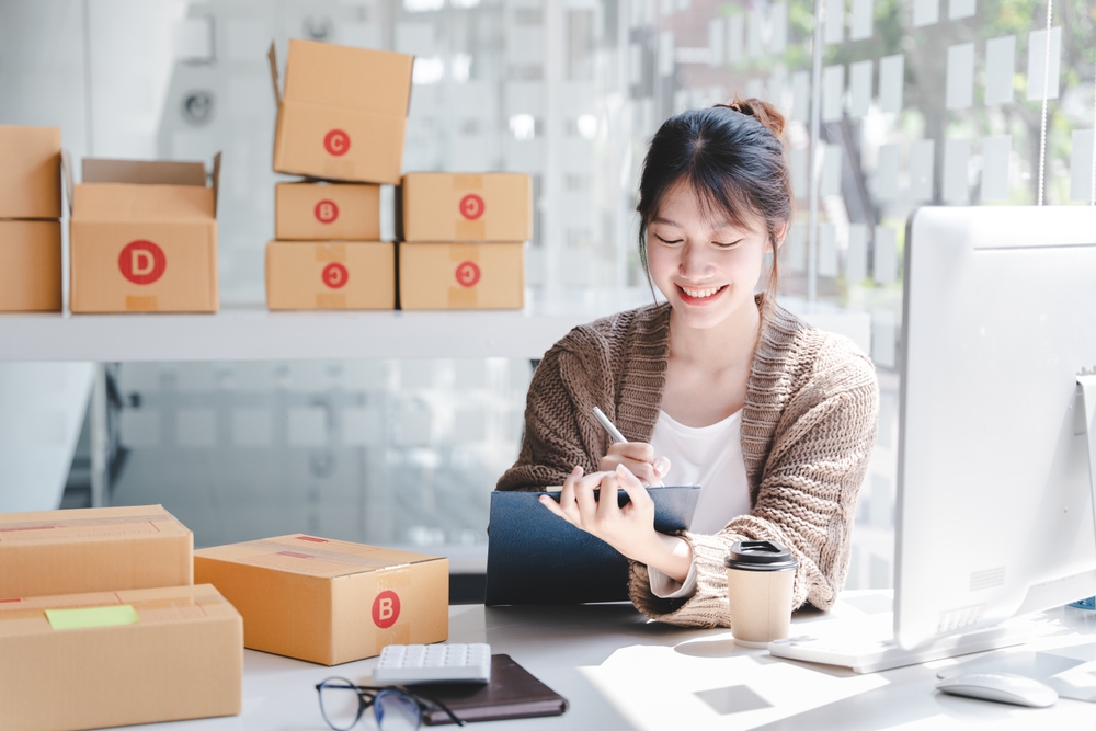 Woman sitting in an office writing notes and smiling surrounded by boxes of inventory