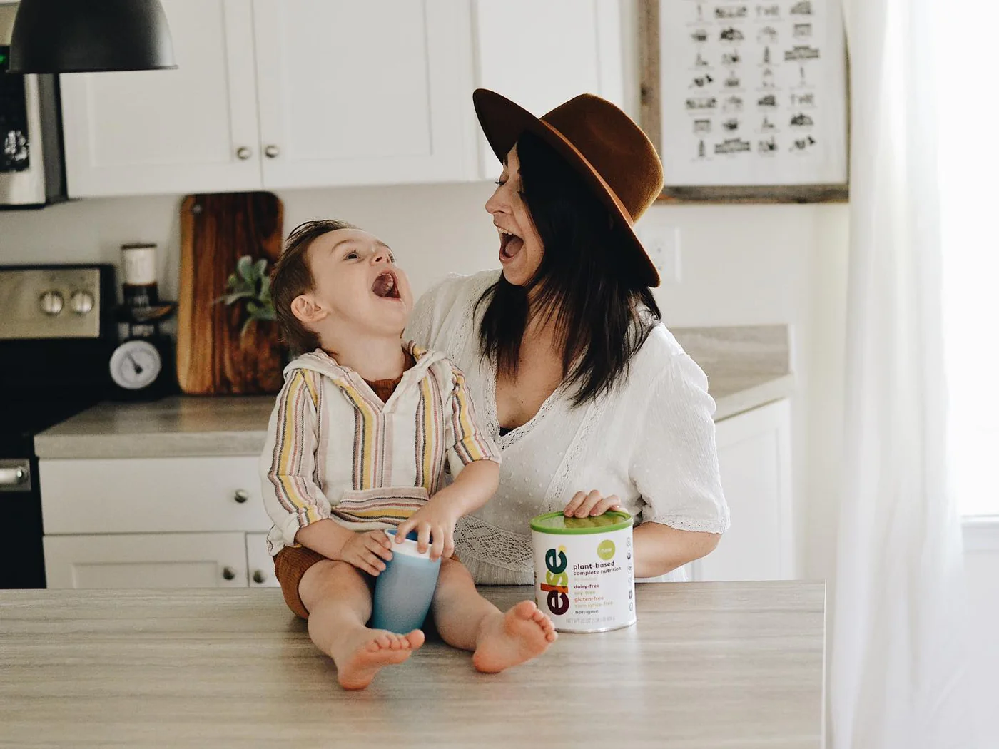 A woman and a child sitting on a kitchen counter with baby formula next to them.