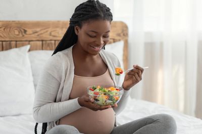 a pregnant woman sitting on a bed eating a salad