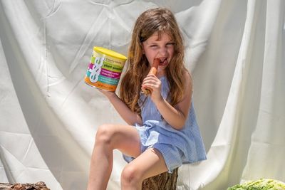 A little girl sitting on a tree stump eating a carrot with formula in her other hand.