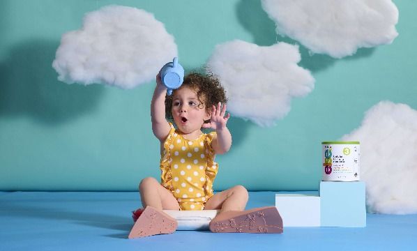 A little girl sitting on the floor next to baby formula with clouds in the background.