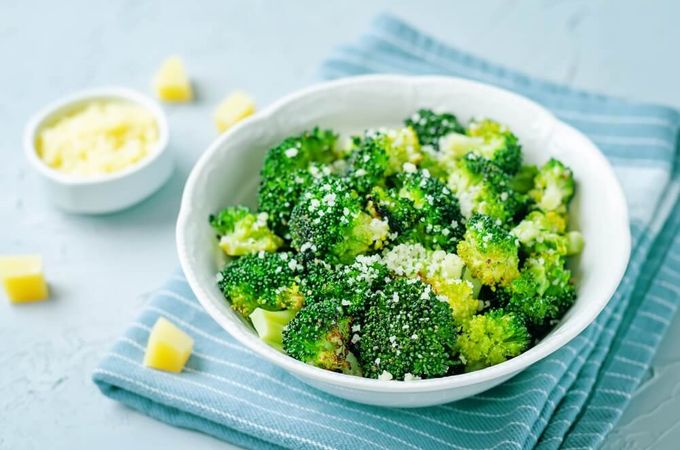 A bowl of broccoli sprinkled with parmesan on a blue cloth and cheese cubes on the side.
