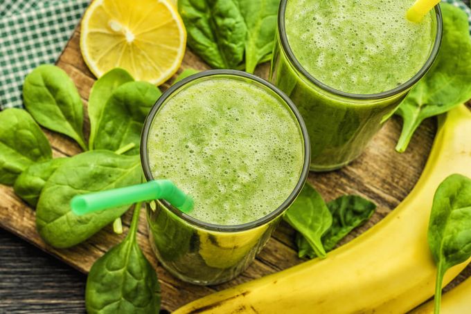 Two glasses of green smoothies next to a banana and some spinach on a cutting board.