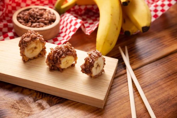 A wooden cutting board topped with banana rolls covered in peanut butter and rice crispies next to fruit and a bowl of cereal.