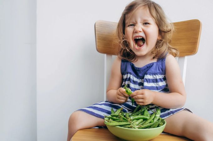 A little girl sitting on a chair with a bowl of green beans.