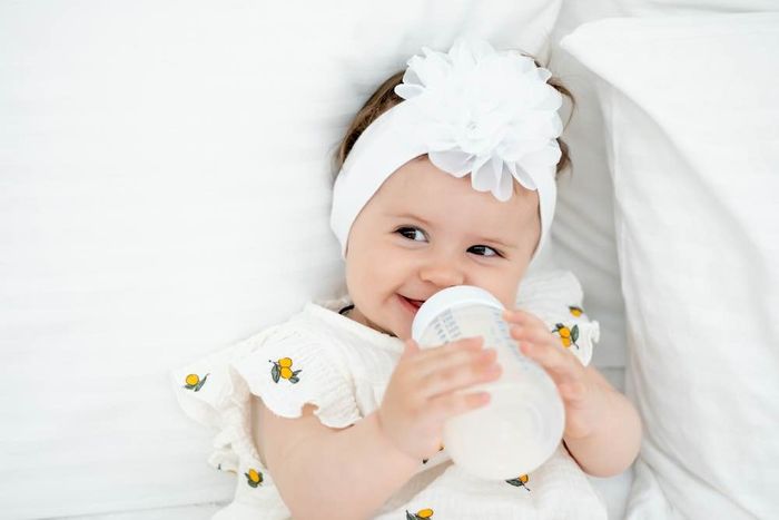 A baby girl drinking from a bottle while laying on a bed.