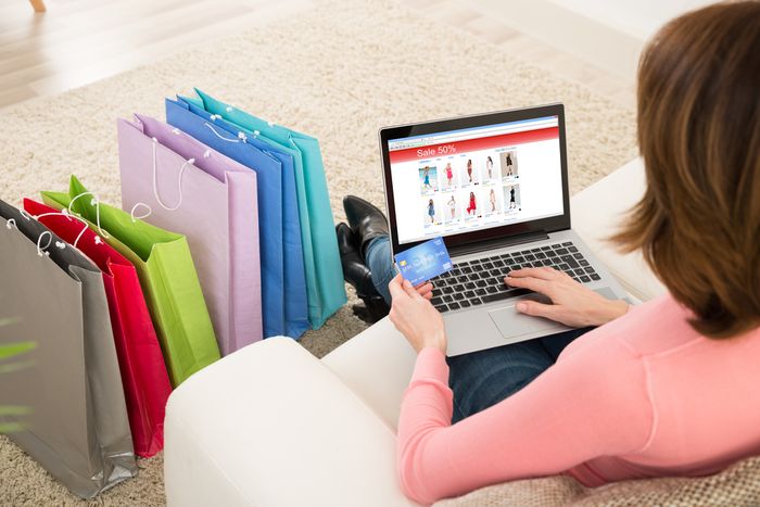 A woman sitting on a couch using a laptop computer to shop online with other shopping bags already arranged next to her feet