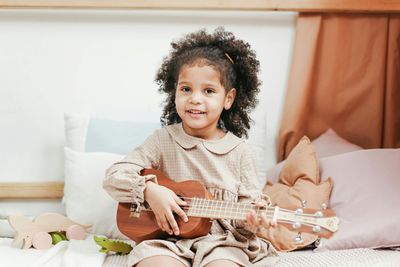 A little girl sitting on a bed playing a ukulele - Best Gold Shoes for Girls to Sparkle 