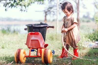 A little girl standing next to a red and yellow wagon, playing with water from a garden hose in her waterproof shoes for kids
