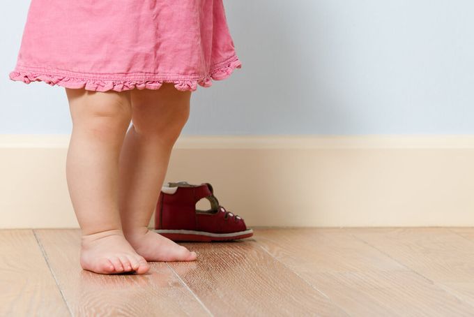 A little girl standing on top of a wooden floor with a red shoe behind her.