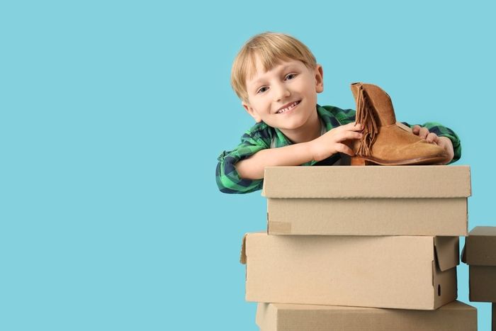 A young boy standing behind a pile of cardboard boxes with a shoe on top.