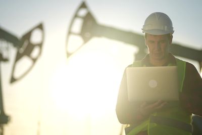 a man in a hardhat using a laptop computer in front of an oil rig