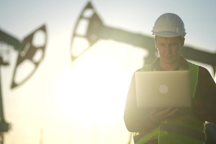 a man in a hardhat using a laptop computer in front of an oil rig