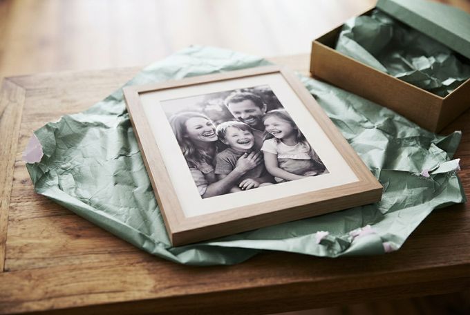 Framed Photo Housewarming Gift Single oak solid-wood frame with a white mat displaying a laughing family, partly wrapped in sage tissue paper.