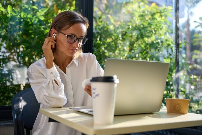 A woman, wearing spectacles and placing wireless earphones in her ear,  seated at small table in a coffee shop, with her laptop and a takeaway coffee cup visible in front of her.